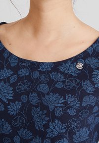 Närbild av en person som bär en marinblå topp med ljusblå blommönster och en liten rund nål på vänster sida nära nyckelbenet.