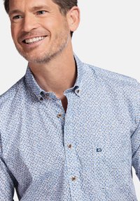 Chemise boutonnière bleu clair avec un petit motif floral répétitif. Tissu texturé et accents de boutons marron. Comprend une poche sur la poitrine.