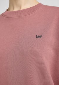 Sudadera de color rosa claro, hecha de una tela suave, con un cuello acanalado y un pequeño logo bordado en negro que dice "Lee" en el pecho.