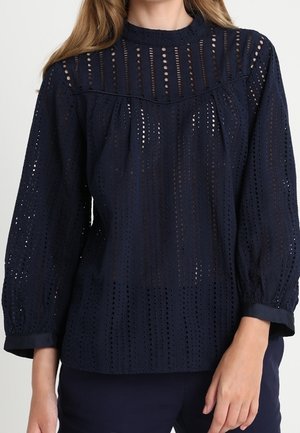 Vrouw draagt een marineblauwe, lange mouw eyelet-blouse met een hoge halslijn en geplooide inzet.