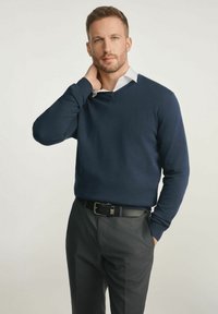 Granatowy sweter w serek wykonany z dzianiny, noszony na białej koszuli z kołnierzykiem. Połączony z szarymi dopasowanymi spodniami i czarnym paskiem.