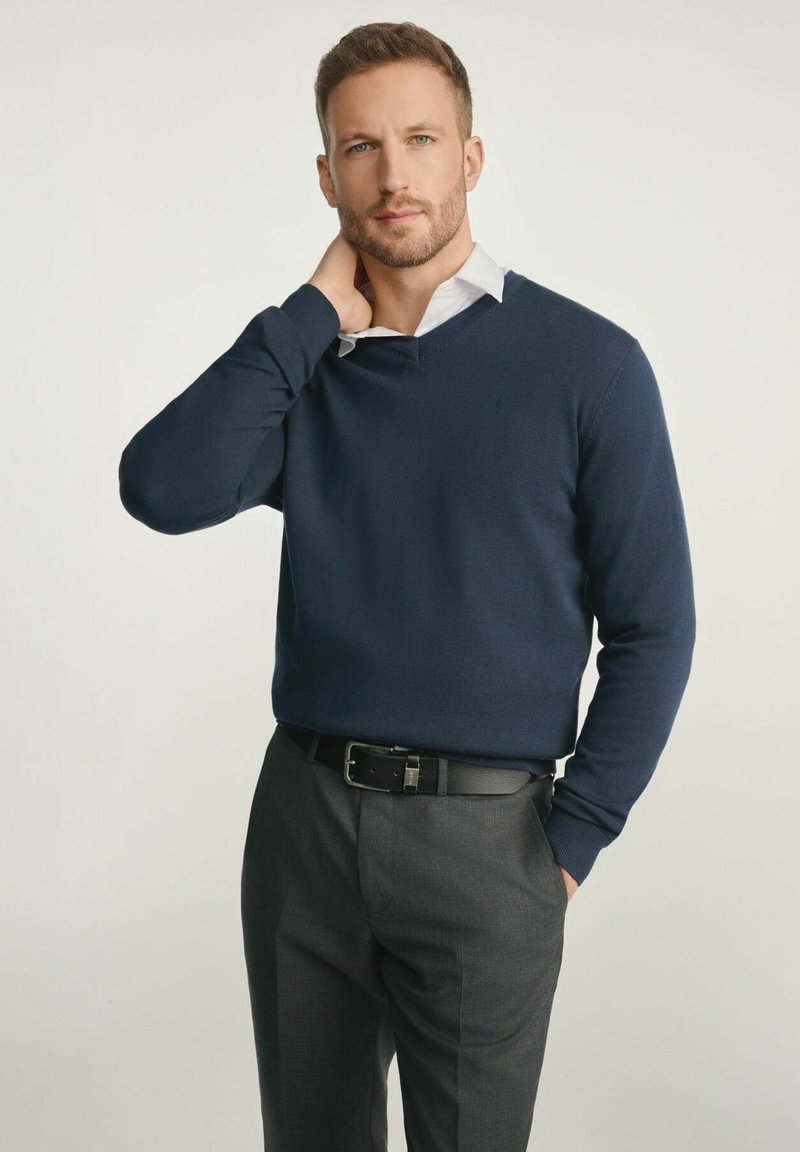 Granatowy sweter w serek wykonany z dzianiny, noszony na białej koszuli z kołnierzykiem. Połączony z szarymi dopasowanymi spodniami i czarnym paskiem.