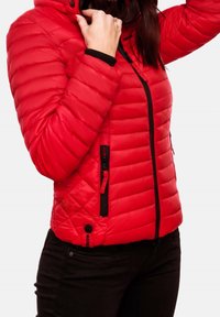 Frau trägt eine rote gesteppte Jacke mit schwarzem Reißverschluss und Taschen, während sie den Kragen anpasst, kombiniert mit schwarzen Hosen.