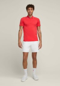 Rotes Poloshirt mit kurzen Ärmeln und einem Viertelreißverschluss sowie weißen Akzenten, kombiniert mit weißen Shorts. Das Modell steht vor einem neutralen Hintergrund.