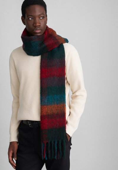 Écharpe en laine avec un motif écossais multicolore dans des tons de rouge, vert et bleu, avec des extrémités à franges, portée sur un pull crème et un pantalon noir.