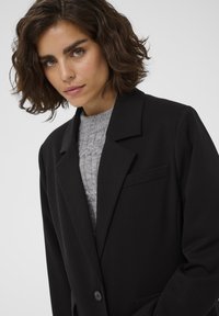 Femme aux cheveux bruns bouclés et courts, portant un blazer noir sur un pull en maille gris, regardant légèrement vers le bas sur un fond uni.