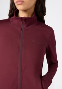 Bordeaux ritsjack gemaakt van gladde, rekbare stof, met een hoge kraag en een subtiele logo op de linker borst.