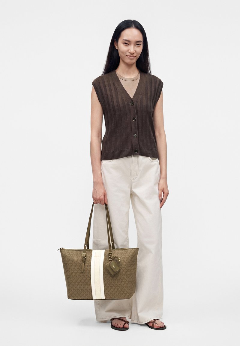 Femme aux longs cheveux foncés porte un gilet marron sans manches, un pantalon blanc, des sandales et tient un grand sac cabas beige avec une bande blanche.