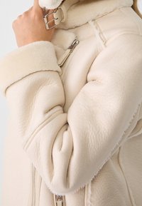 Nahaufnahme eines beigefarbenen Schaffelljackenärmel mit einem gefalteten Bund und einer Hand, die einen silberne Schnalle einstellenden Riemen in der Nähe des Kragens anpasst.