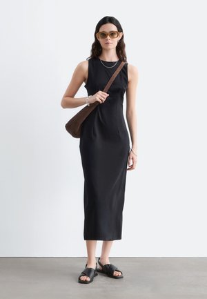 Femme portant une robe midi noire sans manches, sac à épaule marron, sandales noires, lunettes de soleil marron clair et bijoux argentés, debout sur un sol gris.