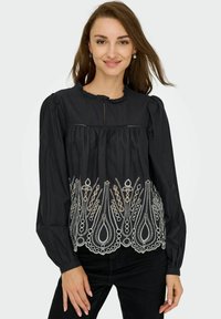 Blouse noire à manches longues avec un design plissé, un col froncé et une broderie blanche complexe à l'ourlet présentant des motifs en forme de goutte.