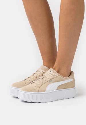 Beżowe zamszowe sneakersy z białymi akcentami, wyściółką wokół kostki oraz grubą białą gumową podeszwą. Widoczne sznurowadła i detal logo po bokach.