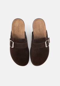 Ζευγάρι σκούρα καφέ σουέντ παπούτσια χωρίς κορδόνια με λεπτομέρεια ασημένιας αγκράφας και μαξιλαρωτές ταμπά εσωτερικές σόλες, φωτισμένα από πάνω σε λευκό φόντο.