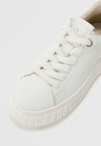 Baskets basses blanches avec une semelle épaisse texturée, des lacets blancs duveteux et un logo subtilement brodé sur la languette, inclinées pour montrer l'avant et le côté.