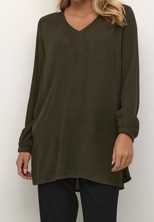 Vrouw draagt een olijfgroene tuniek met lange mouwen en een V-hals, en zwarte broek, staand tegen een effen lichte achtergrond.