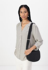 Svart crossover-veske med preget "MOP"-detalj, sammen med en lys beige skjorte med et geometrisk svart mønster.