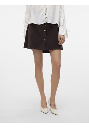 Frau trägt einen dunkelbraunen Rock mit Knopfleiste, weiße Langarmbluse mit Bindebändern und weiße spitze High Heels, steht vor einem einfarbigen Hintergrund.