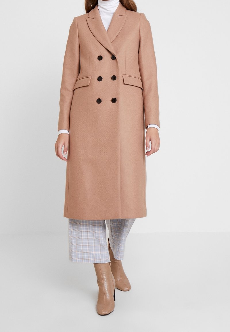 Femme portant un long manteau camel croisé sur un pull à col roulé blanc, un pantalon court à carreaux et des bottines beiges, debout sur un fond uni.