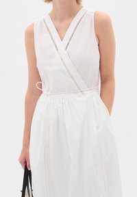 Vestito avvolgente bianco con scollatura a V profonda, design senza maniche e sottili aperture orizzontali lungo il collo. Presenta una cintura da legare in vita.