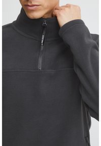 Siv pulover iz flisa z visokim ovratnikom in pol-zadrgo. Ima ravne šive in teksturirano površino, ki poudarja toploto in udobje.