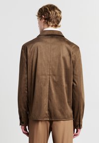 Uomo con capelli biondi mossi che indossa una giacca in suede marrone e pantaloni beige, in piedi di spalle su uno sfondo bianco.