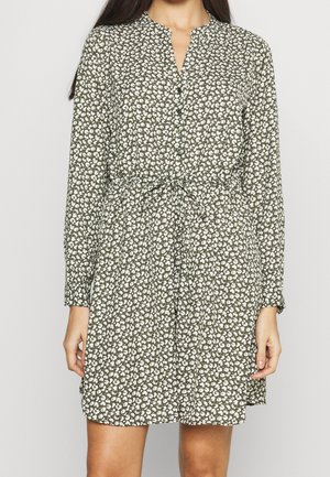 Woman wearing a long-sleeve, olive green dress with small white floral patterns and a tie belt at the waist, standing against a plain background.