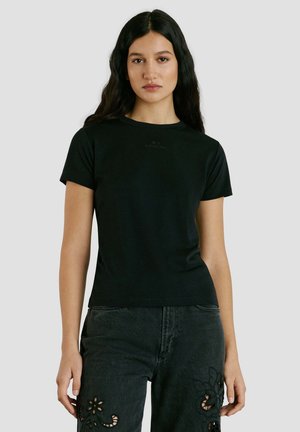 Frau mit langen dunklen Haaren, die ein schlichtes schwarzes Kurzarm-T-Shirt und schwarze Jeans mit floralen Ausschnitt-Designs trägt, steht vor einem grauen Hintergrund.