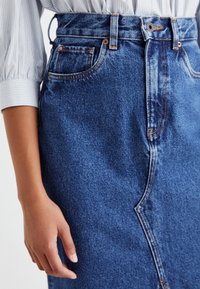 Jupe en denim bleu foncé à taille haute, avec poches avant et des coutures contrastantes. Sa coupe est droite et présente un léger dégradé.