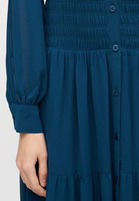 Tealfärgad skjortklänning med en rynkad överdel, långa puffärmar och en kjol i våder med mjuk textur.