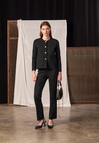 Čierne sako na mieru s zlatými gombíkmi, skombinované s priliehavými nohavicami rovnakej farby a čiernymi lodičkami s ostrou špičkou. Modelka drží malú čiernu kabelku.
