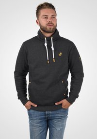 Suéter gris oscuro con cuello alto, cordones blancos, dos bolsillos frontales, detalles amarillos y un logo en el pecho y la manga.