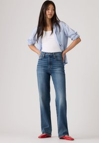 Γυναίκα που στέκεται με τα χέρια στη μέση, φορώντας μπλε πουκάμισο με ρίγες πάνω από λευκό τοπ, τζιν ψηλόμεσα και κόκκινα μυτερά παπούτσια σε απλό φόντο.