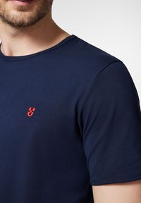 Niebieska bawełniana koszulka z okrągłym dekoltem. Posiada małe, czerwone haftowane logo na lewej piersi. Gładka tekstura, krótkie rękawy.