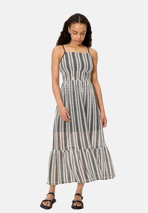 Mujer joven con cabello largo y rizado, vistiendo un vestido midi con estampado en blanco y negro y sandalias negras, de pie con la cabeza ligeramente inclinada.