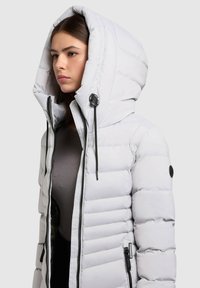 Zimní prošívaná bunda světle šedé barvy s velkou kapucí, předním zipem a černými detaily. Má prošívaný vzor pro izolaci.