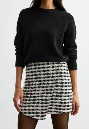 Femme portant un pull noir à manches longues et une jupe portefeuille à carreaux noir et blanc avec des boutons décoratifs.
