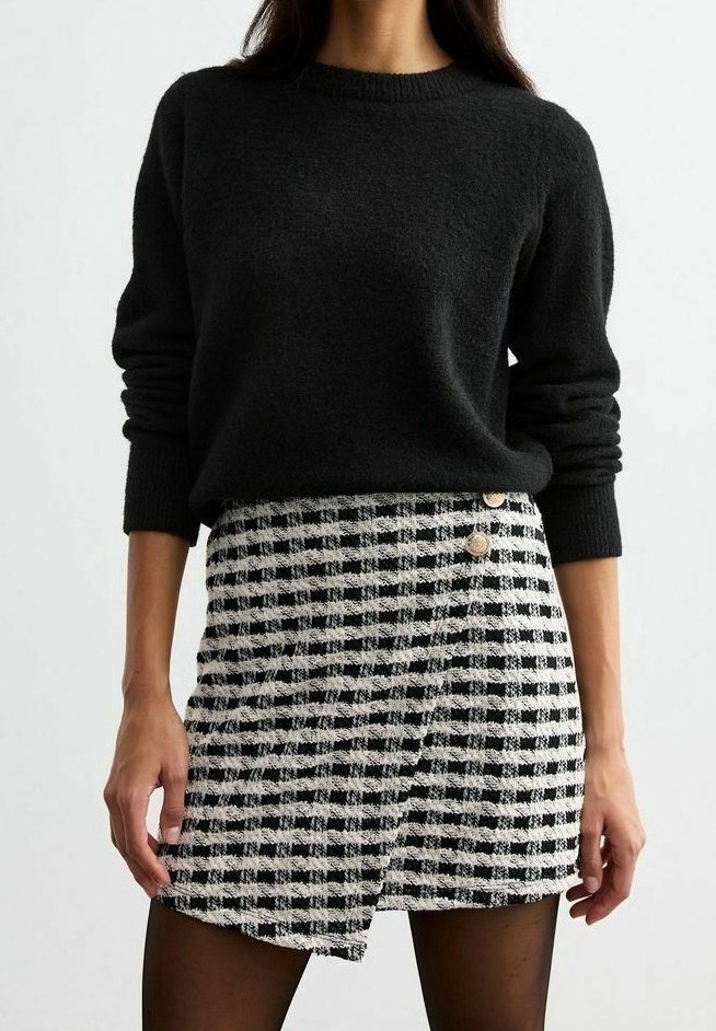 Femme portant un pull noir à manches longues et une jupe portefeuille à carreaux noir et blanc avec des boutons décoratifs.