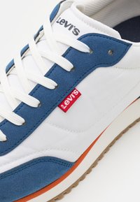 Sneakers marki Levi's z białą tkaniną na cholewce, niebieskimi zamszowymi akcentami, białymi sznurowadłami i czerwoną metką z logo. Gumowa podeszwa z pomarańczowym detalem.