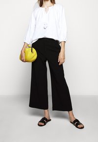 Biała bluzka z bufkami i detalem w kształcie dziurki, połączona z czarnymi spodniami o szerokich nogawkach. Trzyma okrągłą żółtą torebkę o fakturze. Czarne sandały.