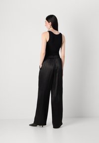 Una modella si trova con le spalle voltate, indossando un top nero senza maniche e pantaloni neri a gamba larga, mostrando un look elegante e moderno.