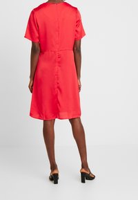 Femme se tenant dos, vêtue d'une robe rouge à manches courtes et longueur genou, ainsi que de sandales noires à talons, sur un fond uni.