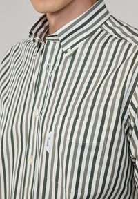 Camisa de botones a rayas con franjas horizontales alternas de color verde oscuro y blanco, cuello entallado y un bolsillo en el pecho con una etiqueta.