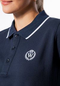 Polo blu navy con bordino bianco, pattina con tre bottoni e logo ricamato che presenta una corona e la lettera "W." Tessuto testurizzato.