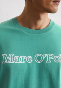 Groene katoen t-shirt met een ronde halslijn en opvallende witte blokletters die "Marc O'Polo" op de borst spellen. Zachte textuur.