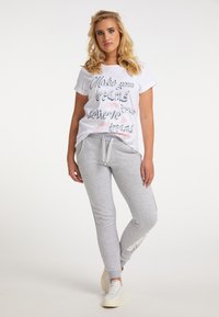 Weißes T-Shirt mit bedrucktem Motivationsslogan; graue Jogginghosen mit seitlichen Akzenten; lässiger Schnitt; strukturierter Baumwollstoff; Modell in stehender Pose.