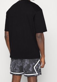 Hombre con camiseta negra de gran tamaño y pantalones cortos de baloncesto negro-gris con un patrón y ribete blanco, de pie contra un fondo claro y liso.