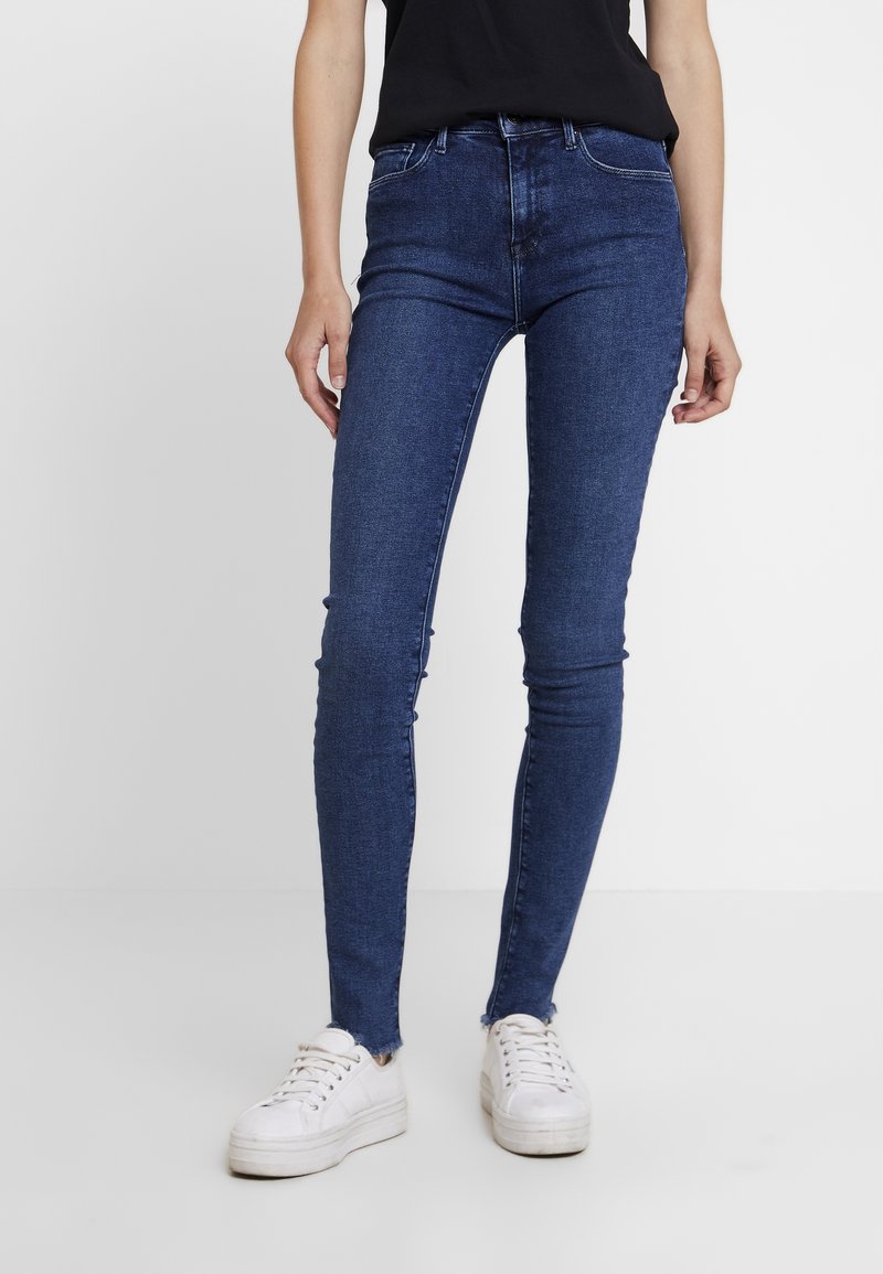 Jeans ajustados en denim azul oscuro con una textura suave, cintura alta y dobladillo deshilachado; combinados con zapatillas blancas para contrastar.