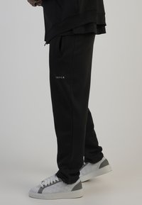 Mustad mustad pingutusvööga püksid, millel on reitel valge logo, koos hallide ja valgete tossudega, millel on tekstuurne disain.