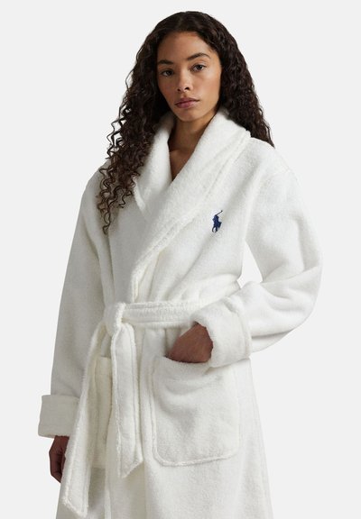 Kvinde med krøllet hår iført en hvid blød badekåbe med sjalskrave, bælte og forlommer, stående mod en ensfarvet baggrund.