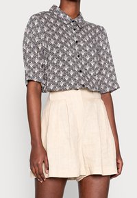 Koszula z krótkim rękawem zapinana na guziki w czarno-białym geometrycznym wzorze, w zestawie z beżowymi lnianymi szortami o wysokim stanie. Miękka tekstura, luźny krój.
